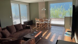 A living room with a brown sofa, a coffee table, and a dining area with a table and chairs. There are large windows with blinds, a wall-mounted light fixture, and a television on a stand. - Guest apartment with breathtaking lake view in the Swiss mountains (Seelisberg)