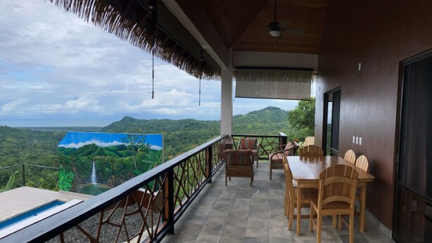 A balcony with a table and chairs, a view of a waterfall, and a thatched roof. - Casa Ceibosito (Tres Rios de Osa)