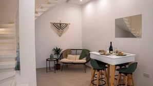 A dining area with a white table, green chairs, and a black chair. There is a plant, a wall hanging, and a staircase in the background.