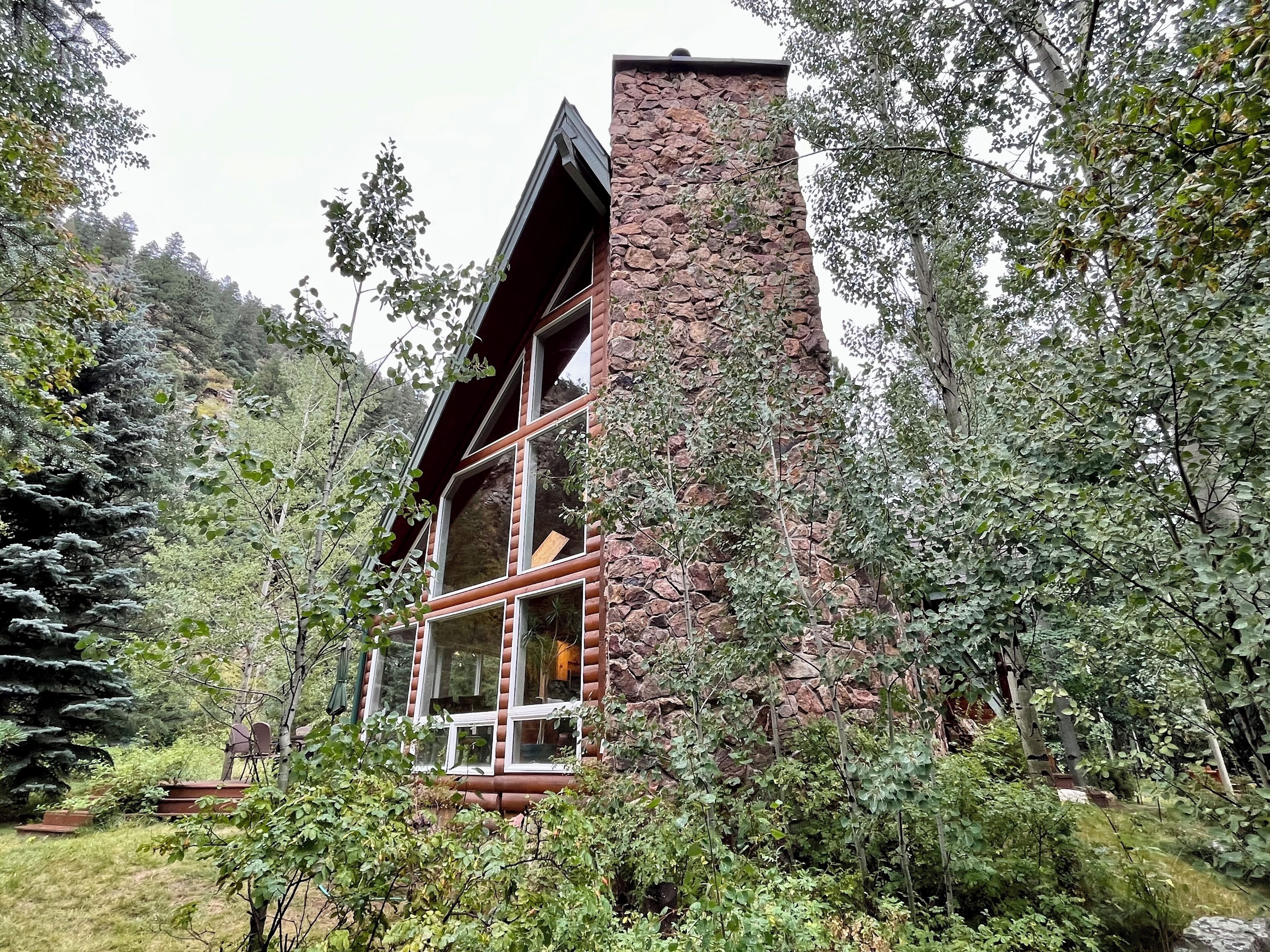 Une cabane en rondins dotée de nombreuses fenêtres et d’une cheminée en pierre, entourée d’une forêt dense.