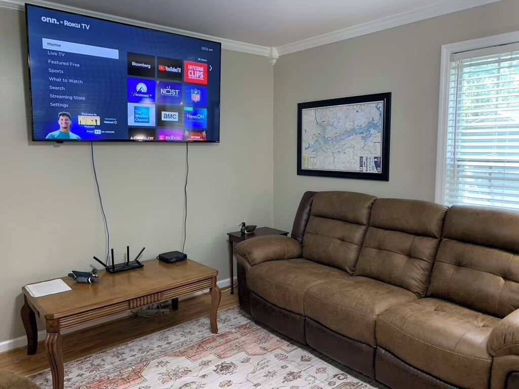Una sala de estar con un televisor de pantalla plana montado en la pared mostrando la interfaz de Roku, un sofá de cuero marrón, una mesa de centro de madera y un mapa enmarcado en la pared.