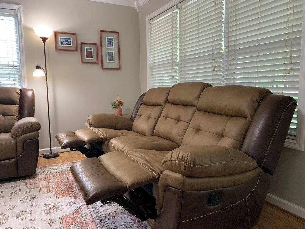 A brown leather recliner with a footrest, positioned in a room with a patterned rug, a floor lamp, and framed pictures on the wall.
