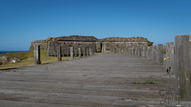 Une promenade en bois mène à des ruines de pierre, sous un ciel dégagé.