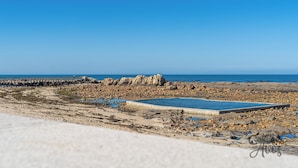 Une plage de galets avec une piscine, une jetée et un ciel bleu azur.