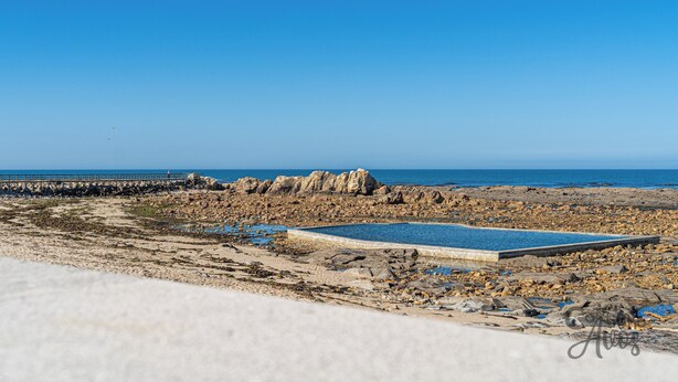 Une plage de galets avec une piscine, une jetée et un ciel bleu azur.