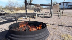 Una chimenea con llamas, rodeada de mobiliario exterior y un edificio al fondo.