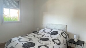 Quarto com cama, mesa de cabeceira, luminária, janela e vista para o verde lá fora.