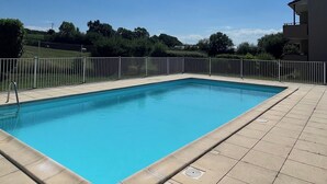 Piscina retangular com deck revestido, cercada por uma vedação branca, e um edifício à direita.