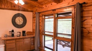 A wooden cabin interior with a kitchen area, a large window, and a wall clock.