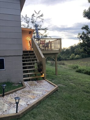 Una casa con terraza de madera, una escalera que lleva a la terraza y un jardín con un sendero e iluminación decorativa.