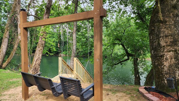Une balançoire en bois avec deux bancs, installée au-dessus d’un petit pont en bois, le tout entouré d’arbres et bordé par un plan d’eau.