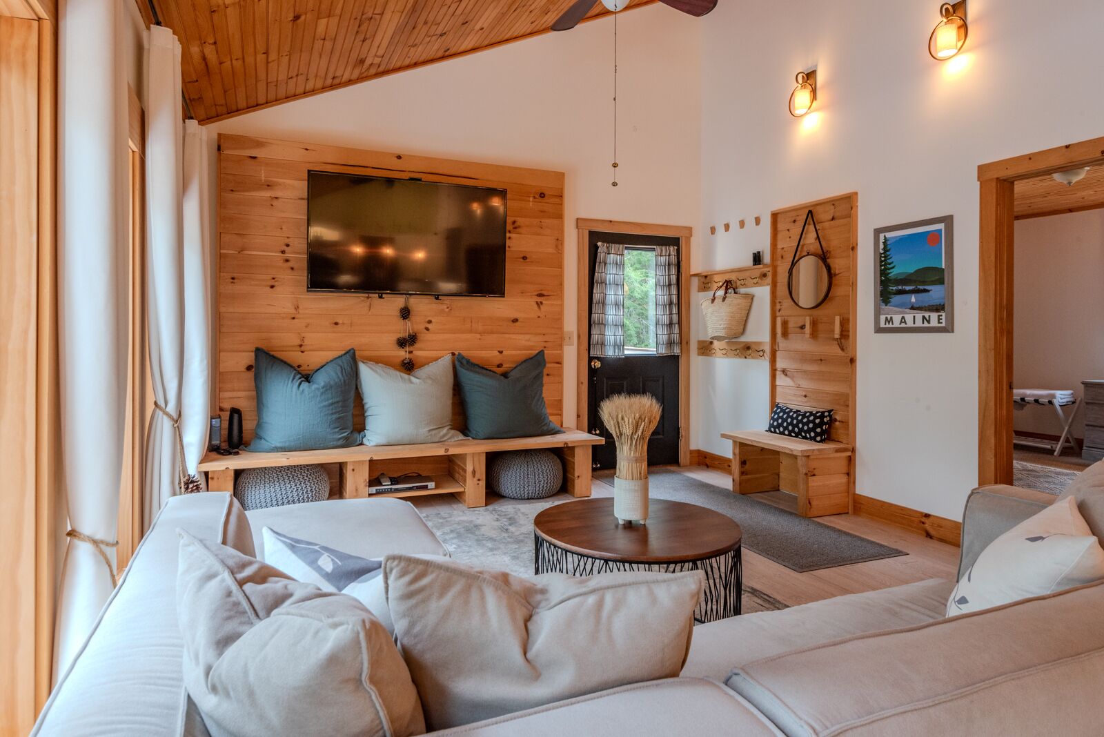 A cozy living room with a large sofa, a coffee table, and a TV mounted on the wall. There is a wooden bench with cushions, a basket on a shelf, and a framed picture of Maine on the wall.