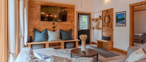 A cozy living room with a large sofa, a coffee table, and a TV mounted on the wall. There is a wooden bench with cushions, a basket on a shelf, and a framed picture of Maine on the wall.