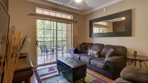A living room with a sofa, ottoman, and a ceiling fan. There is a sliding glass door leading to a balcony with chairs.