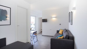 Een moderne woonkamer met een grijze bank, een eettafel met stoelen en een flatscreen televisie aan de muur.