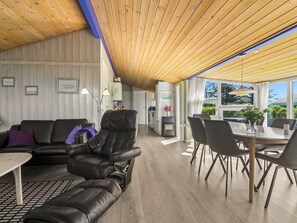 Un interior moderno con sofá de cuero, mesa de comedor y sillas, techo de madera y ventanas amplias.