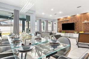 A modern living room with a glass dining table, grey sofas, and a flat-screen TV mounted on a wooden wall. - The Best Home's Resort Orlando (Kissimmee)