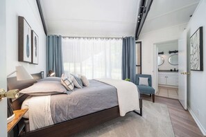 Un dormitorio moderno con cama, mesitas de noche, una silla, un escritorio y un baño con espejo y lavamanos.