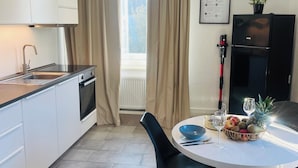 Een moderne keuken met witte kasten, een roestvrijstalen afzuigkap en een zwarte koelkast. Een eettafel met een schaal fruit en een wijnglas staat voor een raam met beige gordijnen.