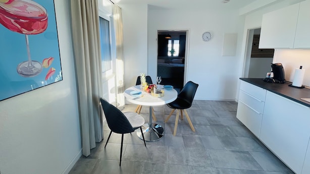 Een moderne keuken met witte kasten, een eethoek met een ronde tafel en stoelen, en een schilderij van een wijnglas aan de muur.