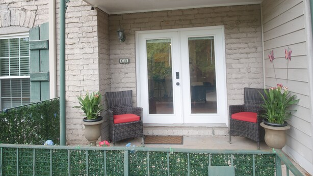 A building entrance with a white door, two wicker chairs with red cushions, and a potted plant on each side.