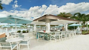 Un espace bar extérieur avec des chaises blanches et des parasols, une piscine en arrière-plan et des palmiers.