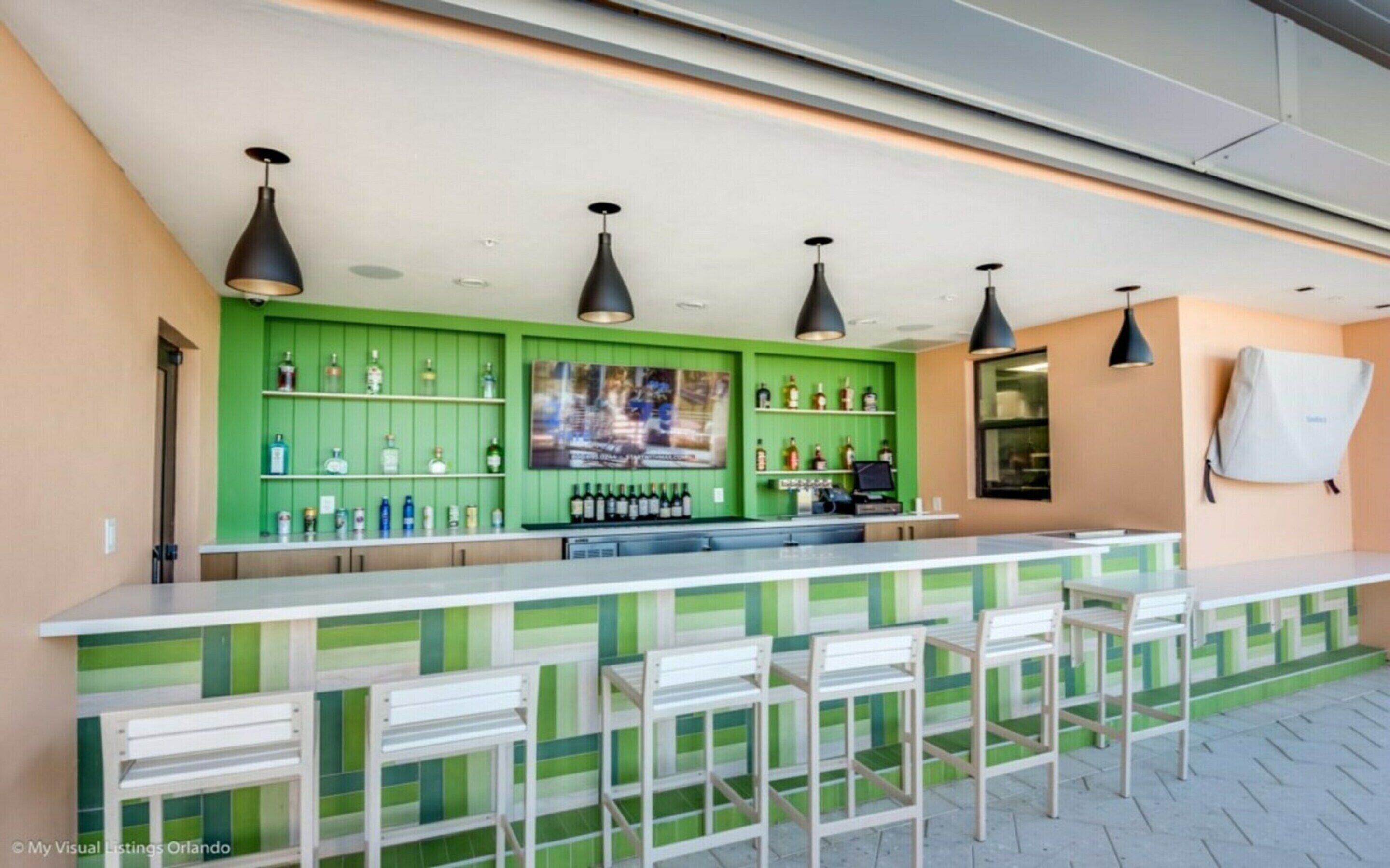 A bar area with green walls, multiple shelves of bottles, and a long counter with chairs.