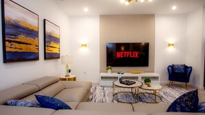 Un salon moderne avec un canapé beige, un téléviseur fixé au mur et deux tables d’appoint ornées d’objets décoratifs.