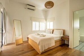 A bedroom with a wooden bed, white bedding, a bedside table, a mirror, and a window with blinds.