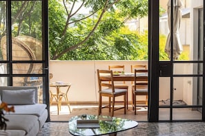 A room with a sofa, a glass coffee table, a dining area with chairs and a table, and a view of greenery through large windows.