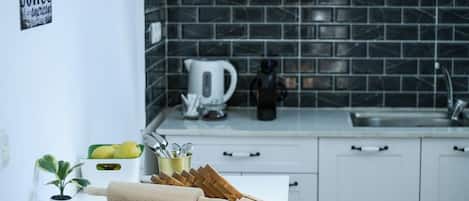 A kitchen with white cabinets, a black tiled backsplash, a kettle, and a cutting board with bread.