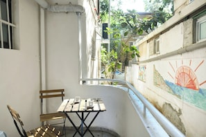 A narrow balcony with a wooden table and chairs, a white wall with a mural, and a view of a street with greenery and buildings.