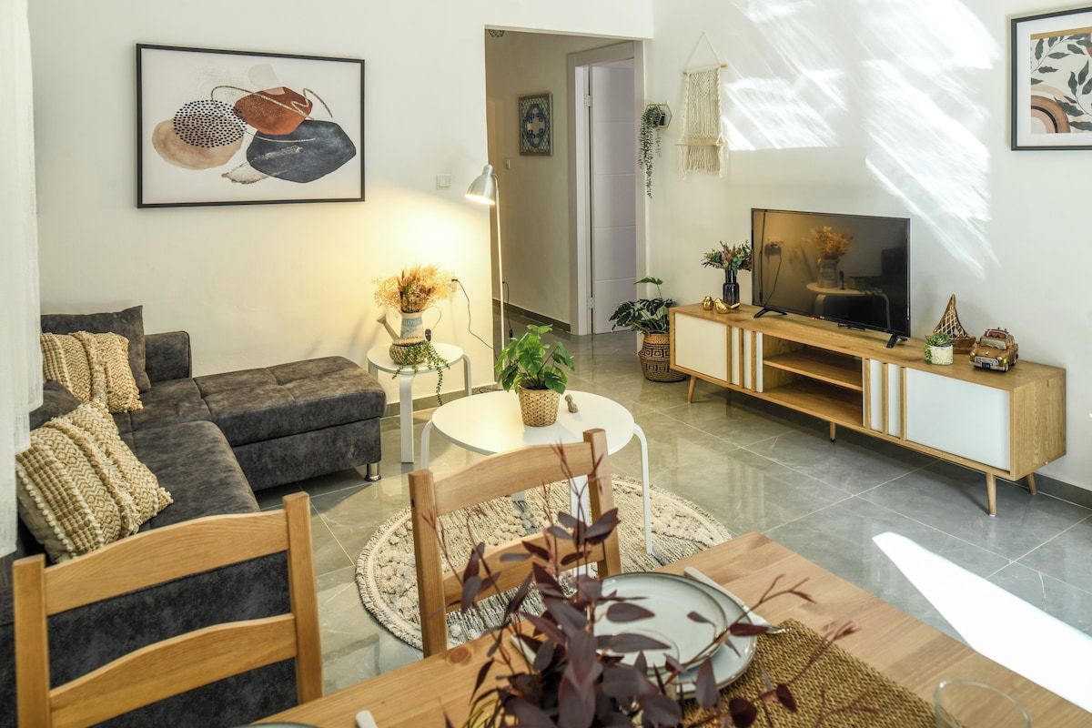 A modern living room with a sofa, a dining table with chairs, a TV on a wooden stand, and decorative plants.