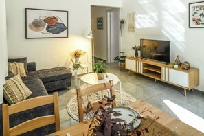 A modern living room with a sofa, a dining table with chairs, a TV on a wooden stand, and decorative plants.