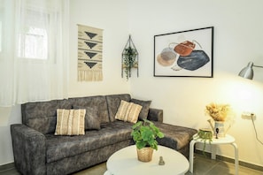 A living room with a grey sofa, patterned cushions, a round white coffee table with a plant, a wall-mounted plant hanger, and a framed abstract artwork.
