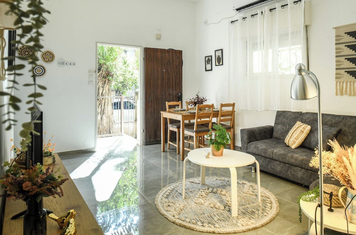 A modern living room with a sofa, a dining table with chairs, a coffee table, and a view of a wooden door leading outside.
