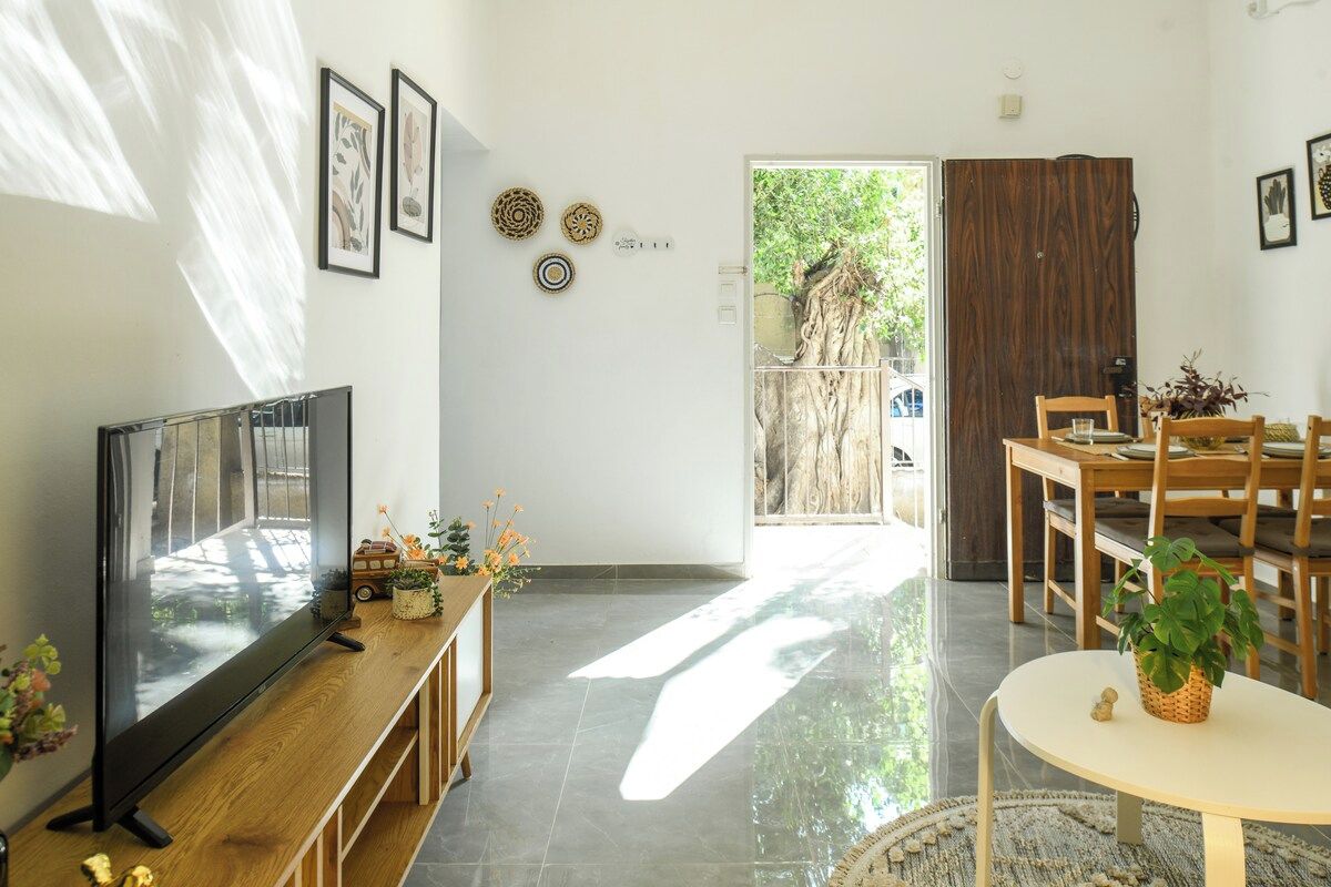A modern living room with a flat-screen TV, wooden furniture, a dining area, and a view of a tree outside.