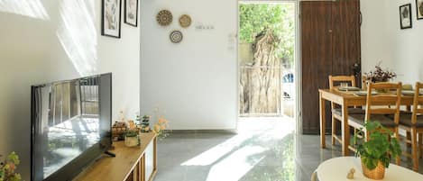 A modern living room with a flat-screen TV, wooden furniture, a dining area, and a view of a tree outside.