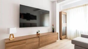 A modern living room with a large flat-screen TV mounted on the wall, a wooden cabinet with drawers, a small table with a lamp, and a potted plant.