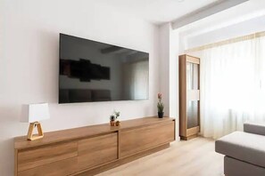 A modern living room with a large flat-screen TV mounted on the wall, a wooden cabinet with drawers, a small table with a lamp, and a potted plant.