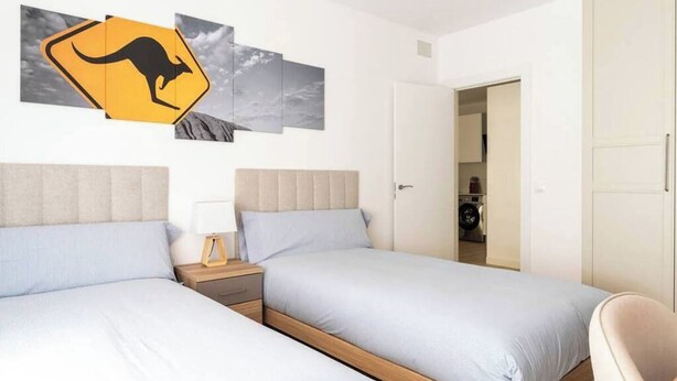 A hotel room with two beds, a nightstand, a wall-mounted kangaroo warning sign, and a view of the sky through the window.