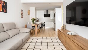 Uma sala de estar moderna com um sofá, uma mesa de jantar com cadeiras, uma área de cozinha com armários e eletrodomésticos, e uma televisão grande fixada na parede.