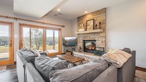 A modern living room with a stone fireplace, a grey sofa, and a wooden coffee table. - Great VIEWS! 10 mins to WISP, boat dock, hot tub, dog friendly, golf course. (Swanton)