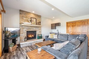 Una sala de estar con chimenea de piedra, sofá gris, mesa de centro de madera y televisor.