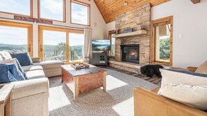 A cozy living room with a stone fireplace, a television, and a large window overlooking a scenic view. - Great VIEWS! 10 mins to WISP, boat dock, hot tub, dog friendly, golf course. (Swanton)