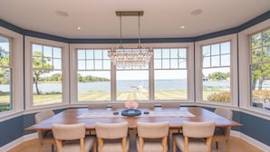 Une salle à manger avec une grande table et six chaises, de grandes fenêtres donnant sur un lac et un quai, et un lustre suspendu au plafond.