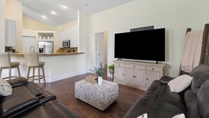 A modern living room with a dark leather sofa, a white ottoman, and a flat-screen TV on a wooden stand. A kitchen with white cabinets and a dining area with bar stools is visible in the background. - Jupiter's Beacon Villa. Less than 2 miles from the beach in the heart of Jupiter (Jupiter)