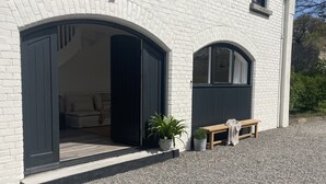 Edifício de tijolos brancos com uma grande porta em arco que dá para o interior. Há um banco de madeira com um cobertor ao lado da porta, e duas plantas em vasos colocadas do lado de fora.