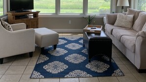 Una sala de estar con ventilador de techo, un televisor sobre un mueble de madera, un sofá color beige, un tapete azul estampado y ventanas grandes con persianas.