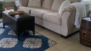 Una sala de estar con un sofá color beige, un otoman de cuero oscuro, un tapete estampado y un barril de madera.